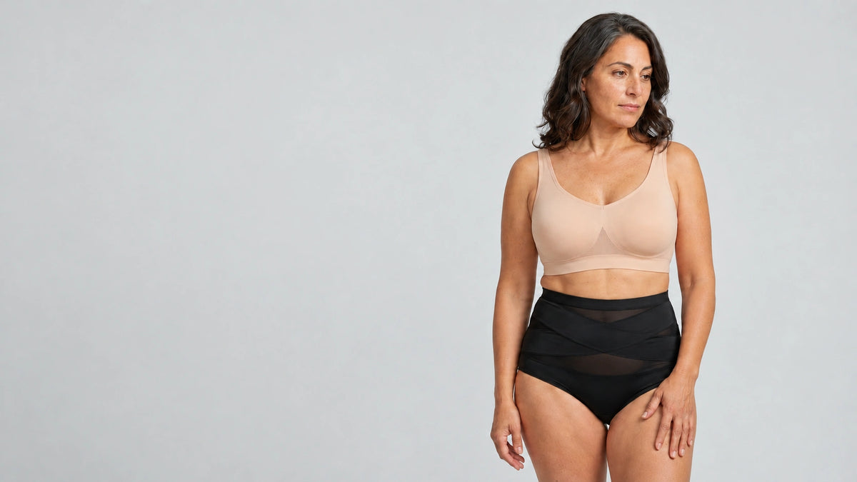 A woman modeling a beige wireless bra and black high-waisted briefs against a light gray studio background.